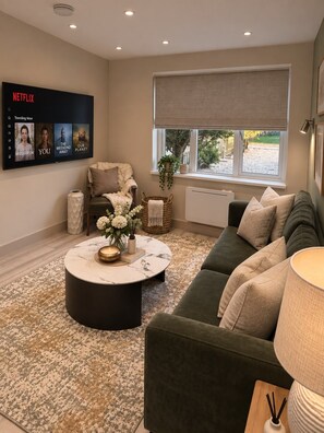 A living room with a green sofa, a round coffee table with flowers, a TV displaying Netflix content, and a window with a view of the outdoors. - Burton House|4 Bed|Sleeps8|Private Parking (Wool)