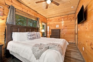A wooden cabin room with a bed, a ceiling fan, a television, and a window with blinds. - Cozy Cabin Near Dollywood & Pigeon Forge w/ Hot Tub (Sevierville)