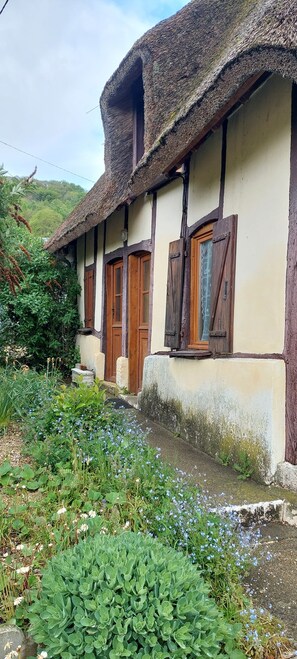 A traditional building with a thatched roof, wooden doors, and a small garden area with various plants. - Country Cottage - A Quiet Norman Thatched Cottage Close to Nature (Marais-Vernier)