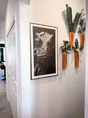 A hallway with a framed artwork of a creature wearing glasses, wooden vases with plants, and a white door. - Luxury Golf Penthouse Verdanza (Ave Maria)