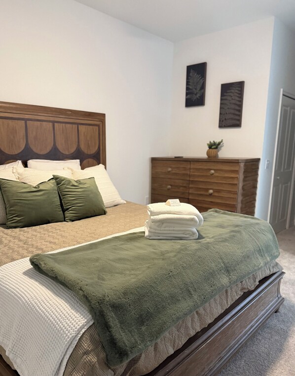 A bedroom with a bed, pillows, a green blanket, a wooden dresser, and two wall art pieces.