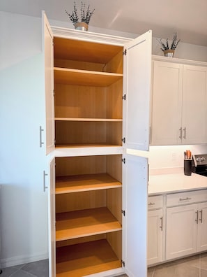 Una cocina con armarios blancos, una despensa de doble puerta con varios estantes y una encimera con un portalapapeles.