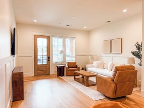A spacious living room with a wooden floor, a white sofa, a brown leather chair, a coffee table, and a TV mounted on the wall. - Homewood Luxury 2 Bedroom 2.5 Bath Townhome 3018 (Homewood)
