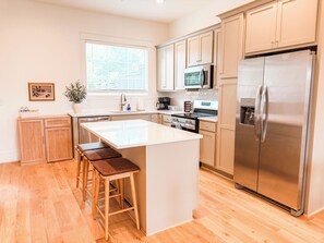 A modern kitchen with wooden cabinets, a stainless steel refrigerator, a central island with stools, and a window providing natural light. - Homewood Luxury 2 Bedroom 2.5 Bath Townhome 3018 (Homewood)