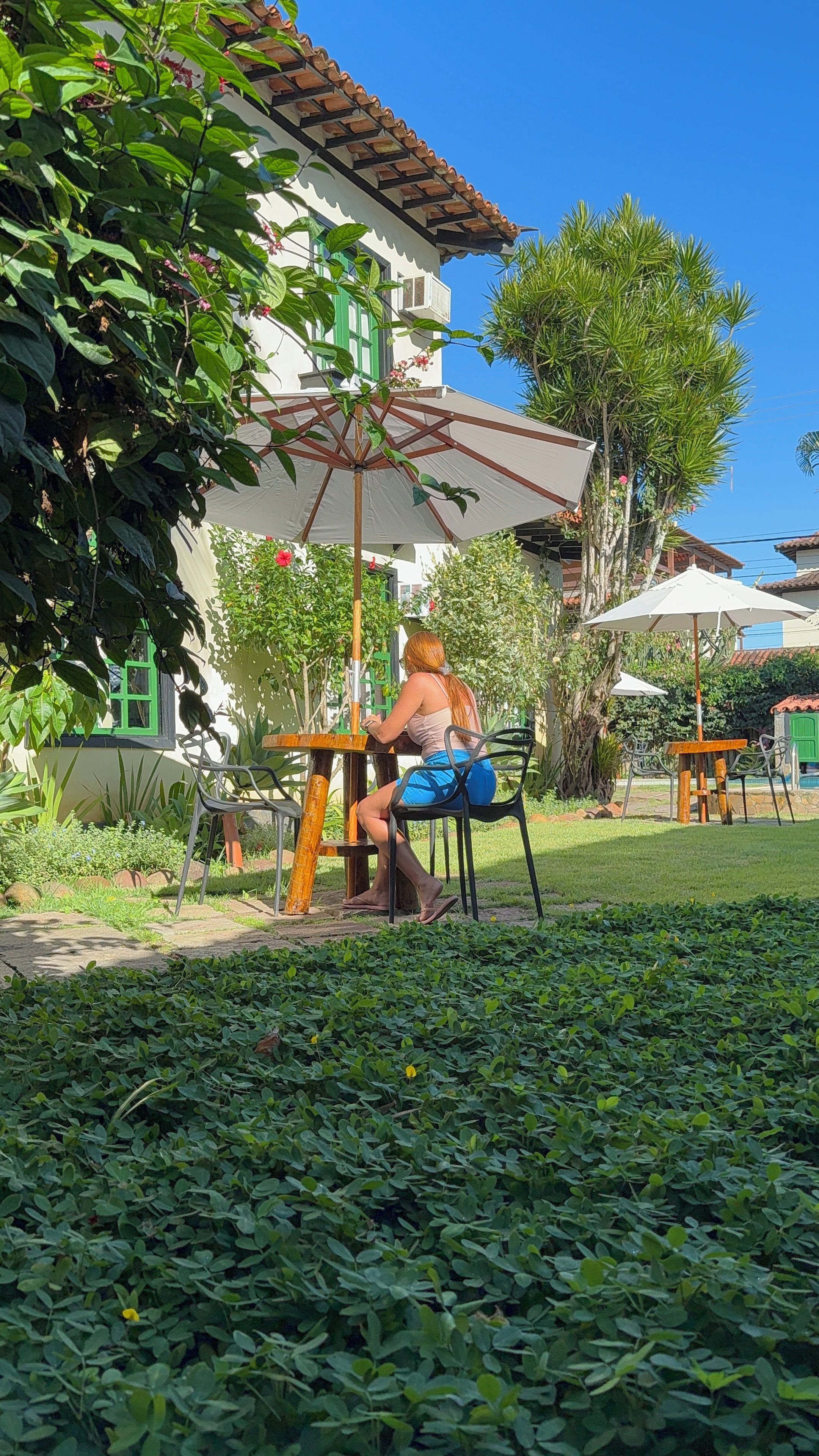 A person seated at an outdoor table with a white umbrella, surrounded by greenery and a building with a tiled roof.