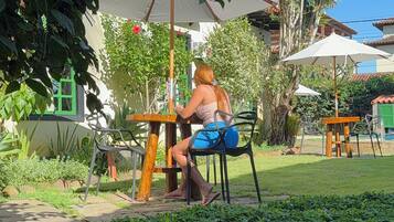 A person seated at an outdoor table with a white umbrella, surrounded by greenery and a building with a tiled roof.