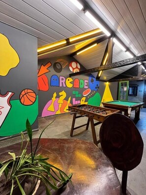 A billiards room with a pool table, a foosball table, and a colorful mural on the wall. - Pousada Sonho Verde (Rio das Ostras)