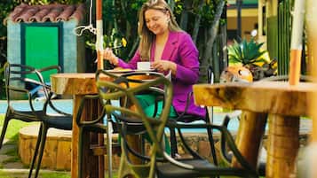 An outdoor dining area with tables and chairs, surrounded by greenery and a building with a green door.