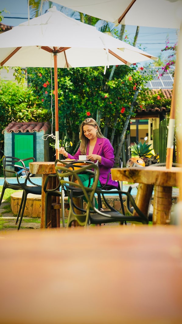 An outdoor dining area with tables and chairs, surrounded by greenery and a building with a green door.