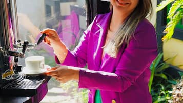 A person in a purple jacket operating a coffee machine, with a window view and a table with a pitcher and cups.