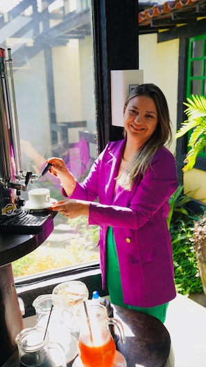 Una persona con una chaqueta morada operando una máquina de café, con vistas al exterior y una mesa con una jarrón y tazas.