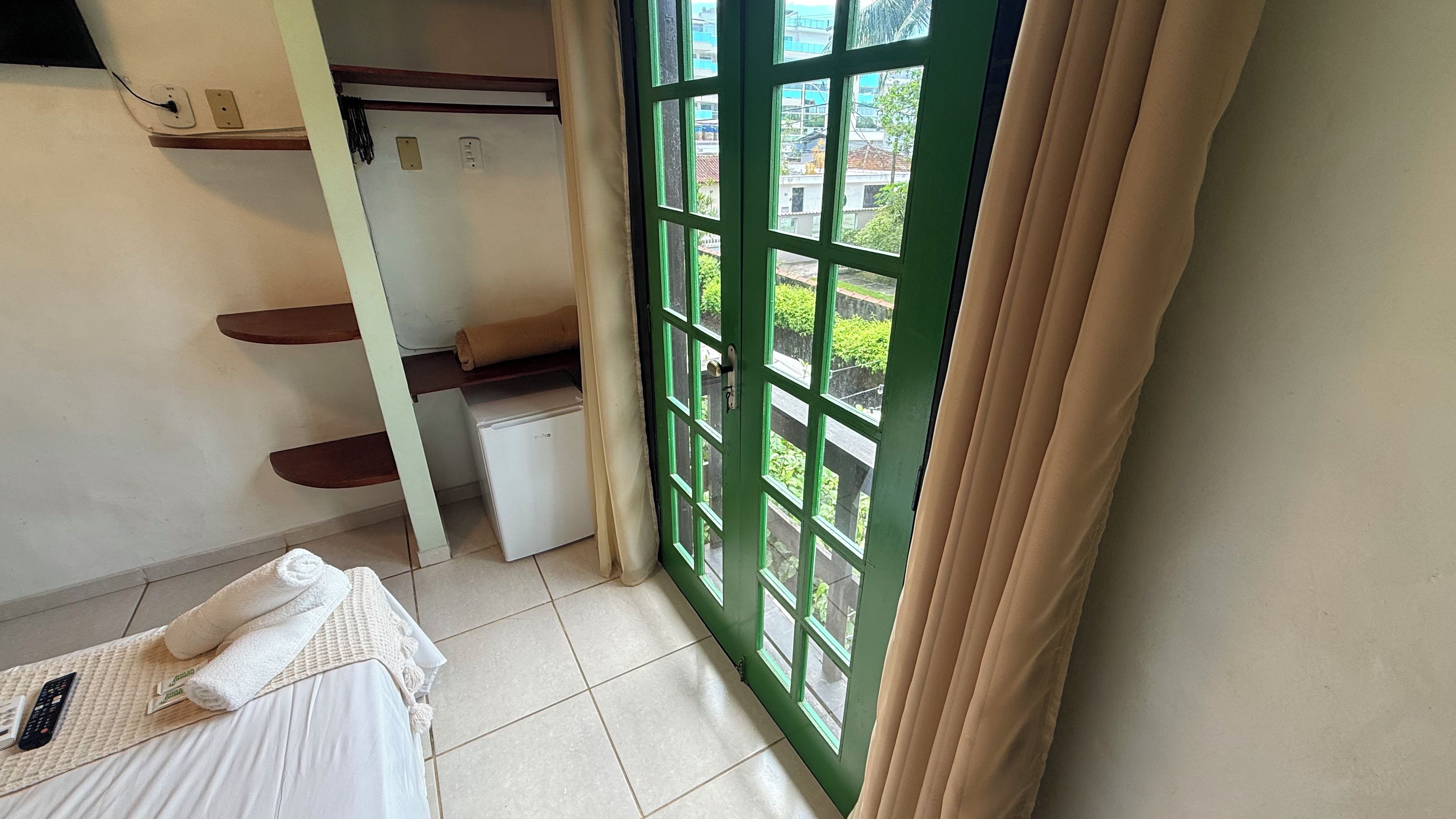 A hotel room with a bed, a small refrigerator, a window with curtains, and a view of greenery outside.