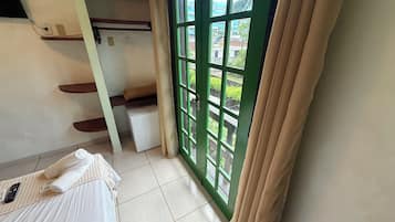 A hotel room with a bed, a small refrigerator, a window with curtains, and a view of greenery outside.