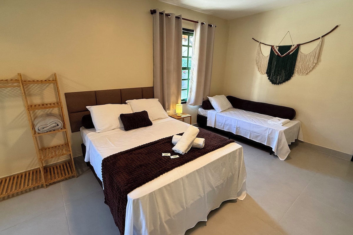 Habitación de hotel con una cama grande, otra más pequeña, un armario, una ventana con cortinas y un adorno de macramé en la pared.