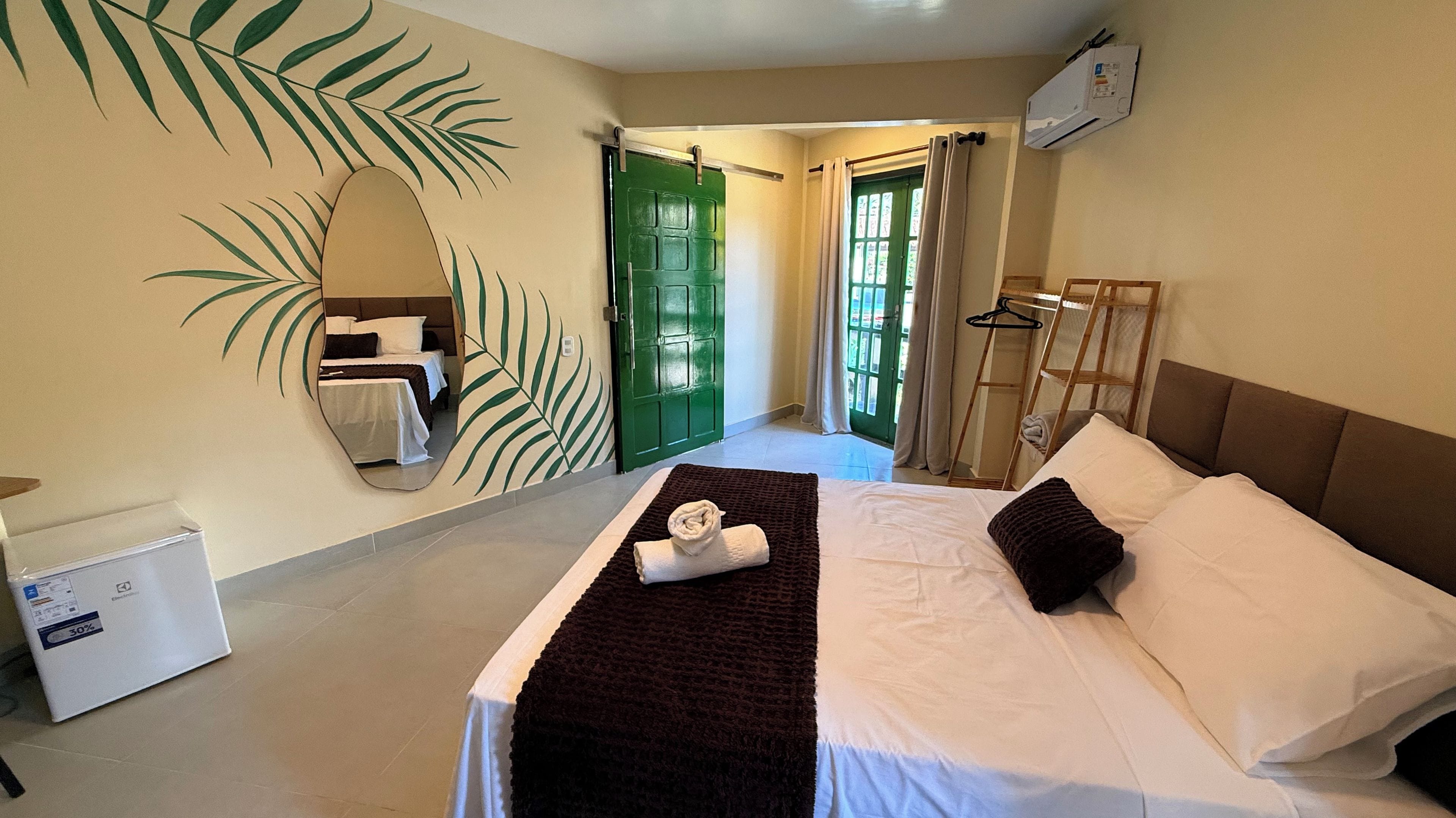 A hotel room with a bed, a mirror, a green door, and a view of the outside.