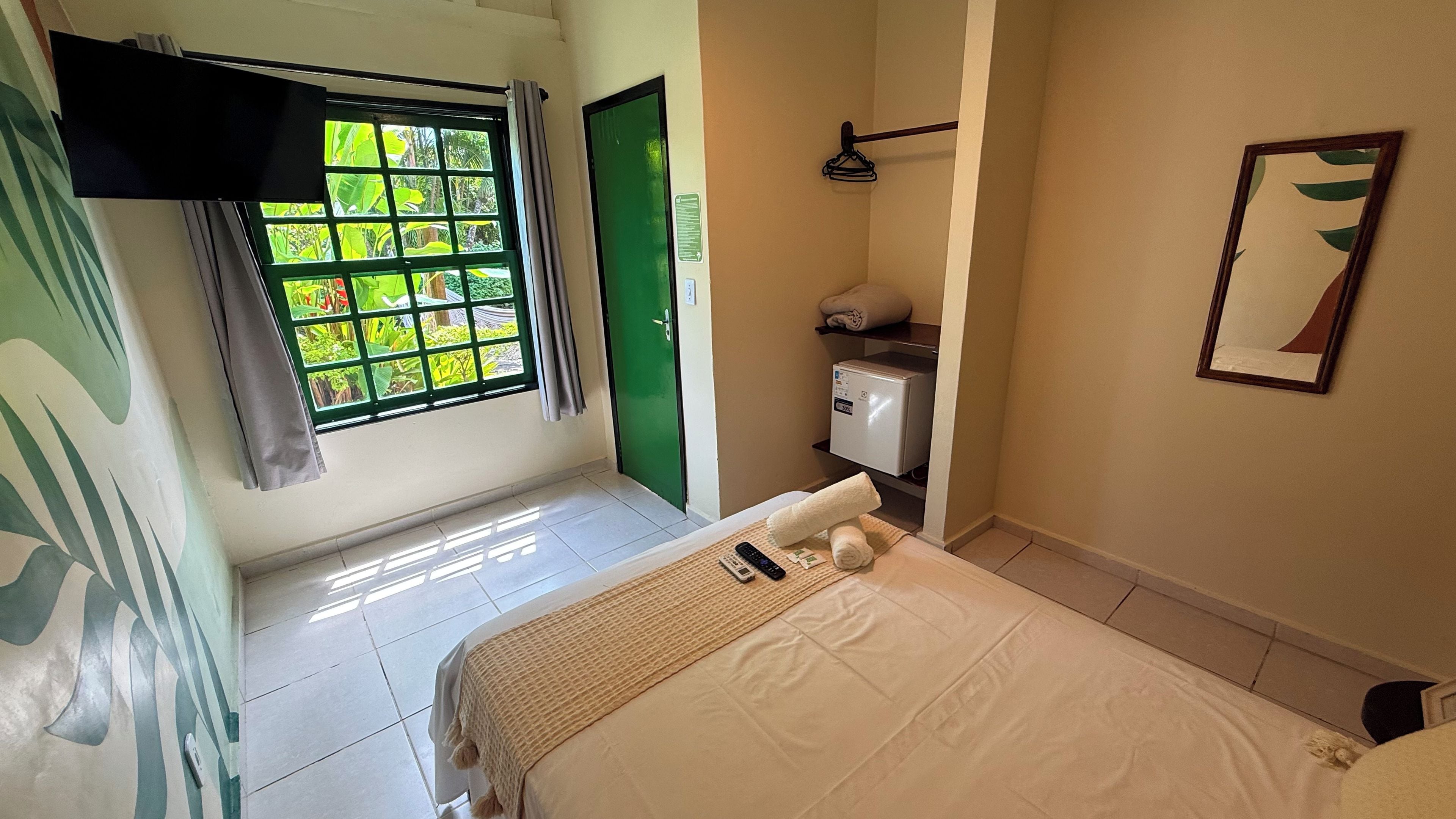 A hotel room with a bed, a small desk, a green door, a window with curtains, and a wall-mounted towel rack.