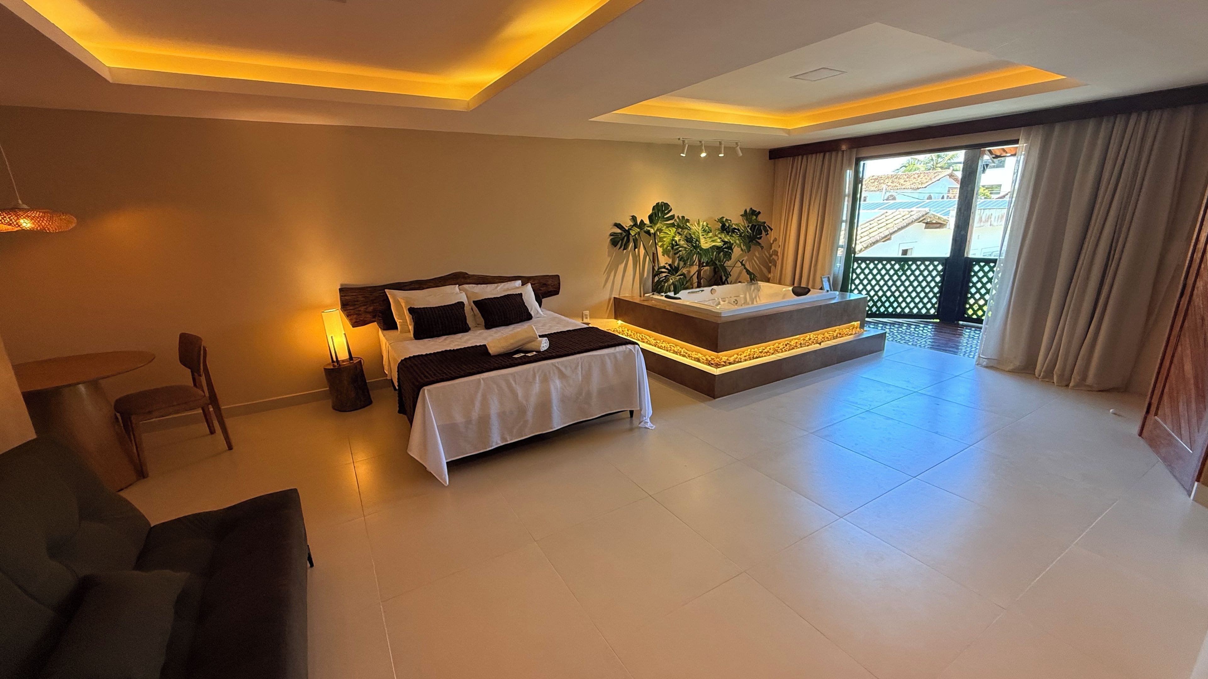 A modern hotel room with a bed, a small table with chairs, a bathtub, and a view of the outside.