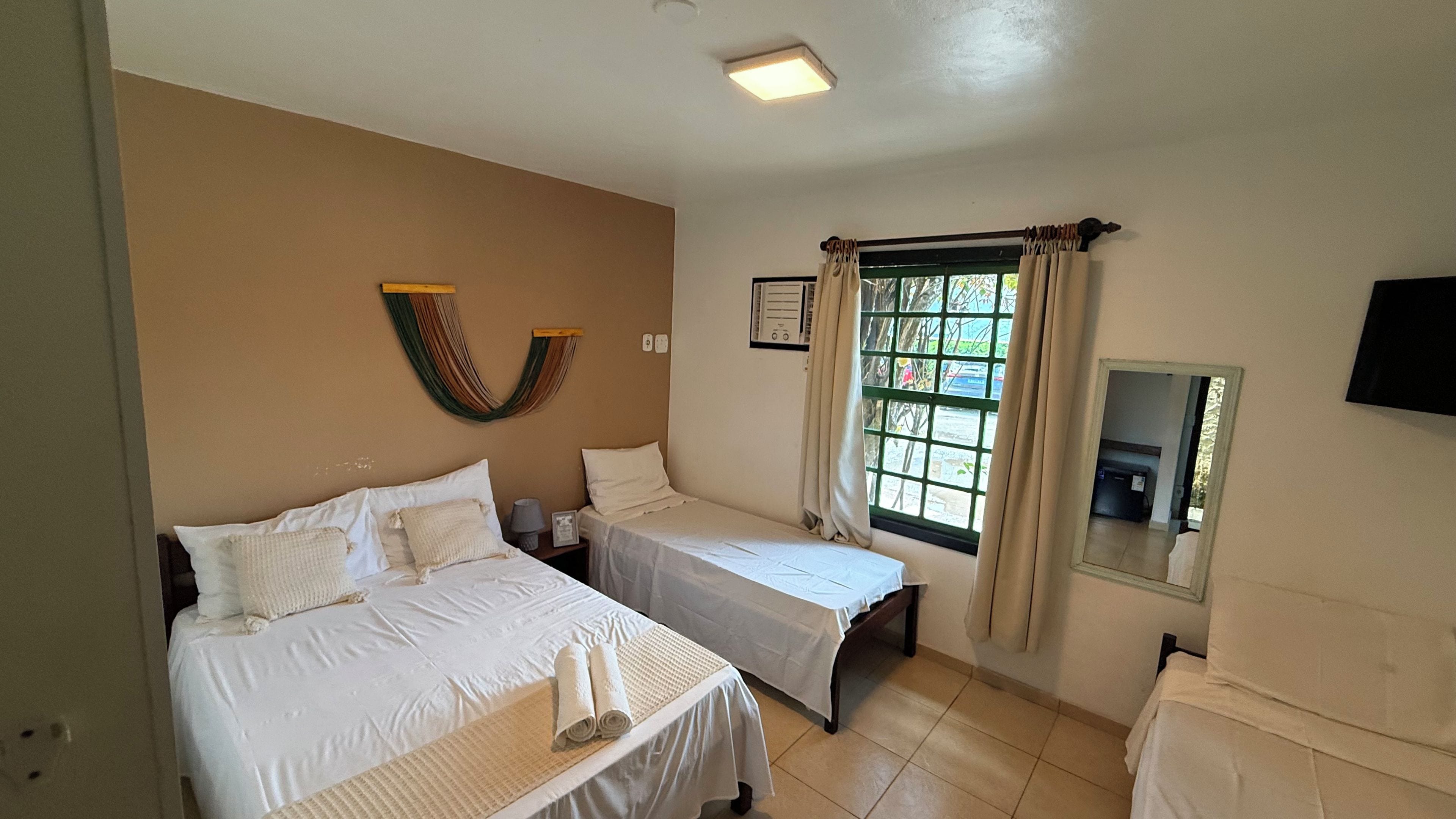 A hotel room with two beds, a window with curtains, a flat-screen TV, and a wall-mounted heater.