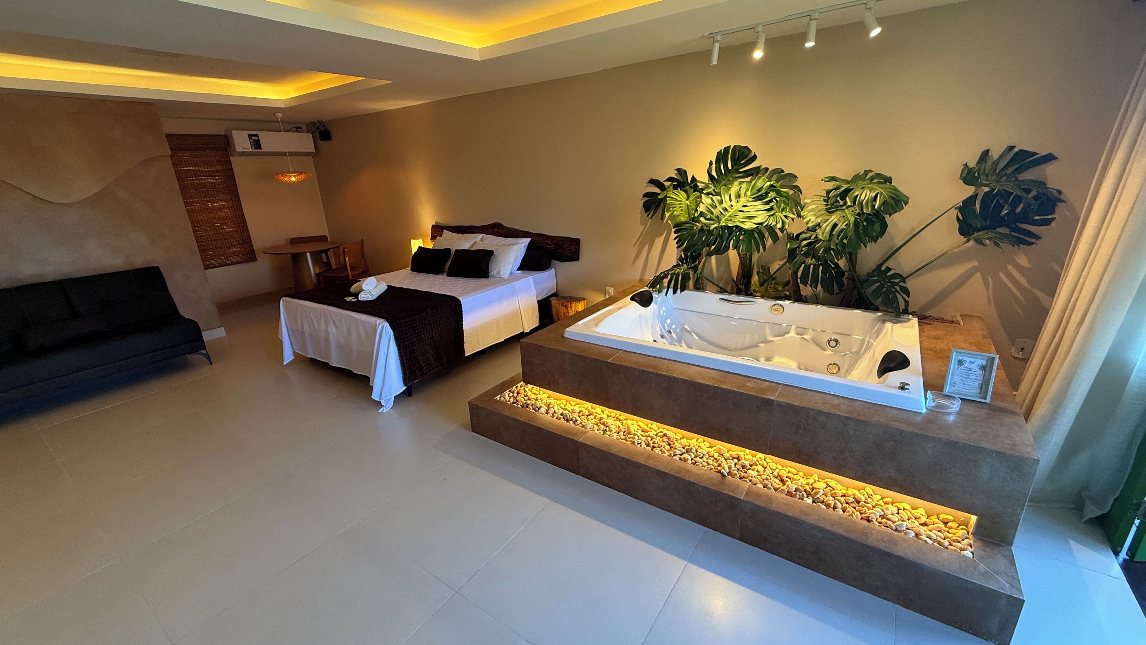 A modern hotel room with a bed, a sofa, a jacuzzi, and large potted plants.