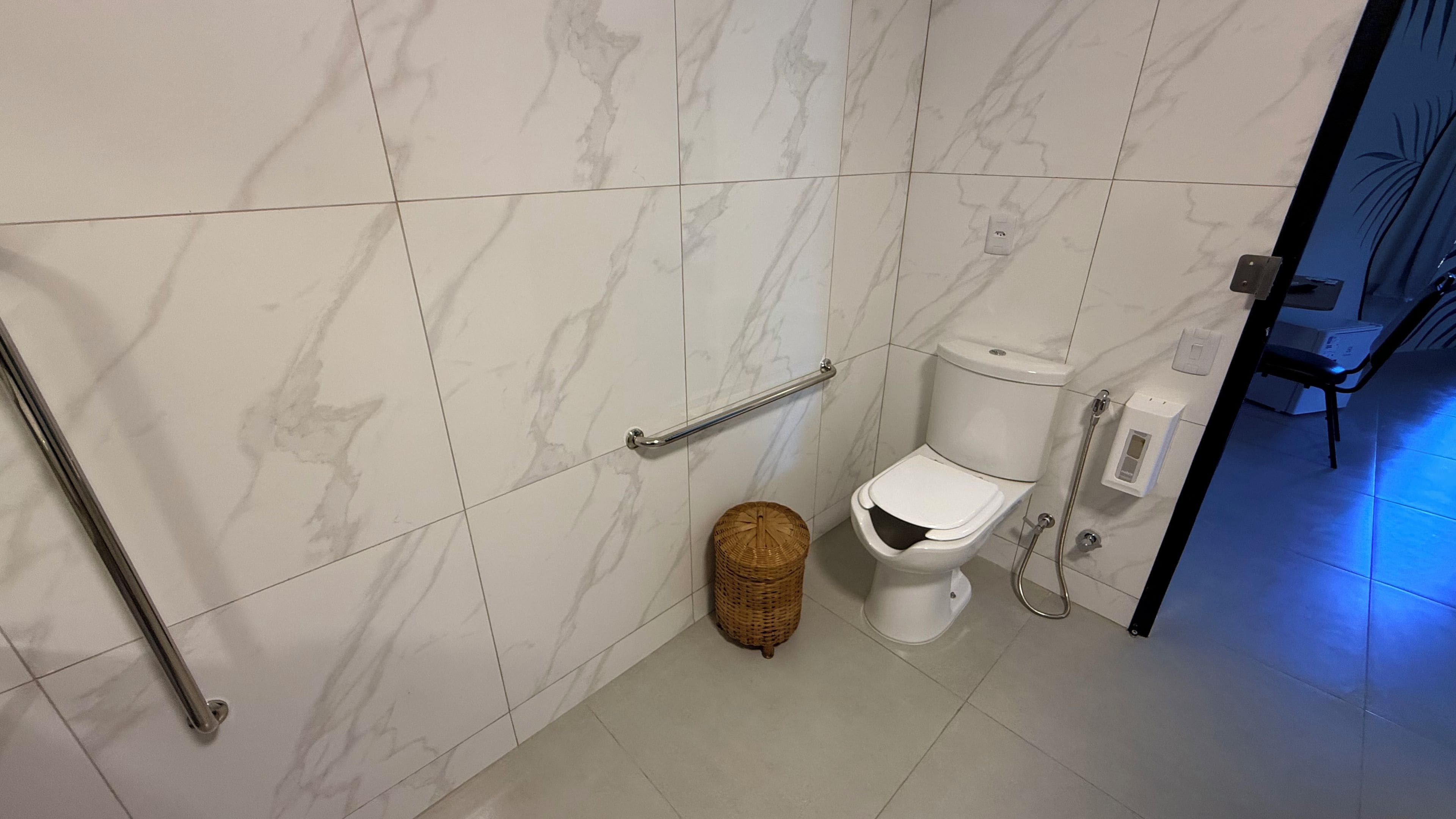 A bathroom with a toilet, a handrail, a waste basket, and a shower area.