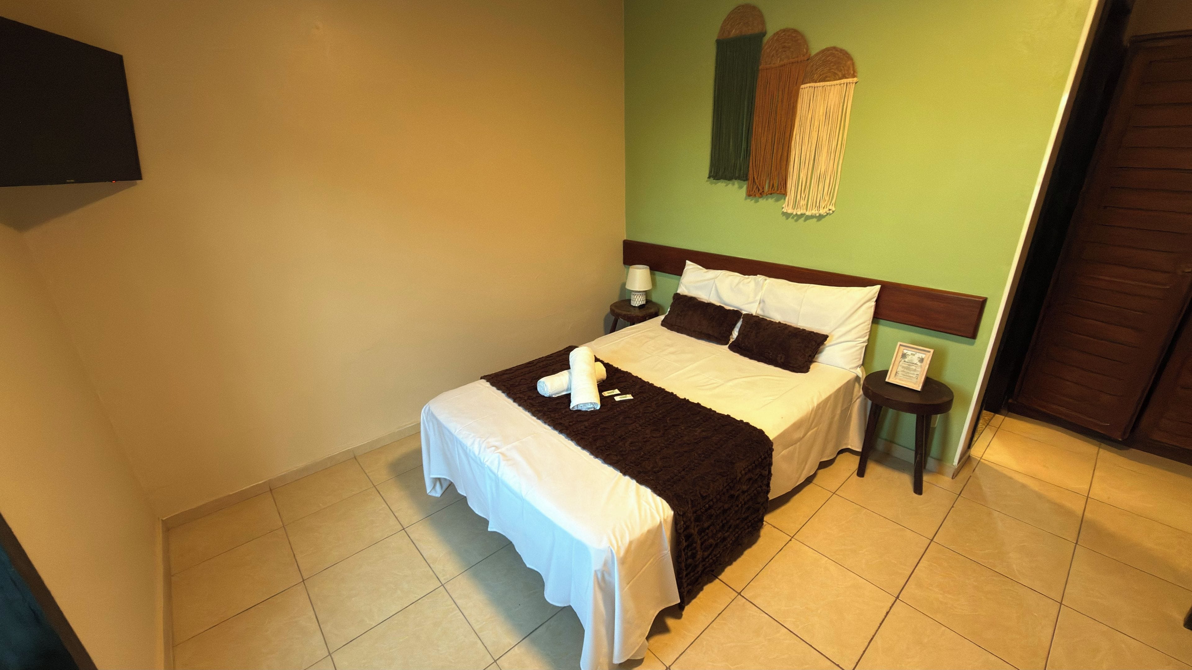 Una cama bien hecha con sábanas blancas y un plaid marrón, una mesita de noche con lámpara, y un cuadro en la pared.