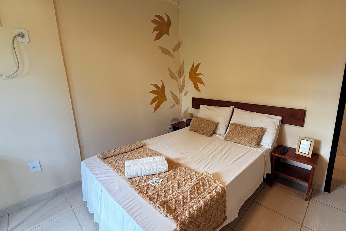 A hotel room with a bed, a nightstand, a wall-mounted power outlet, and a wall decoration.