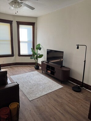 A living room with a flat-screen TV, a floor lamp, a potted plant, and a large rug. - Quaint 2-bedroom house in Detroit perfect for your getaway (Detroit)
