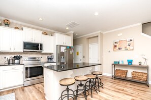 A modern kitchen with white cabinets, stainless steel appliances, a black countertop, and wooden stools. - Crab House - Oceanfront w Pool, Hot Tub, Elevator & Backyard. Amazing Views! (North Topsail Beach)