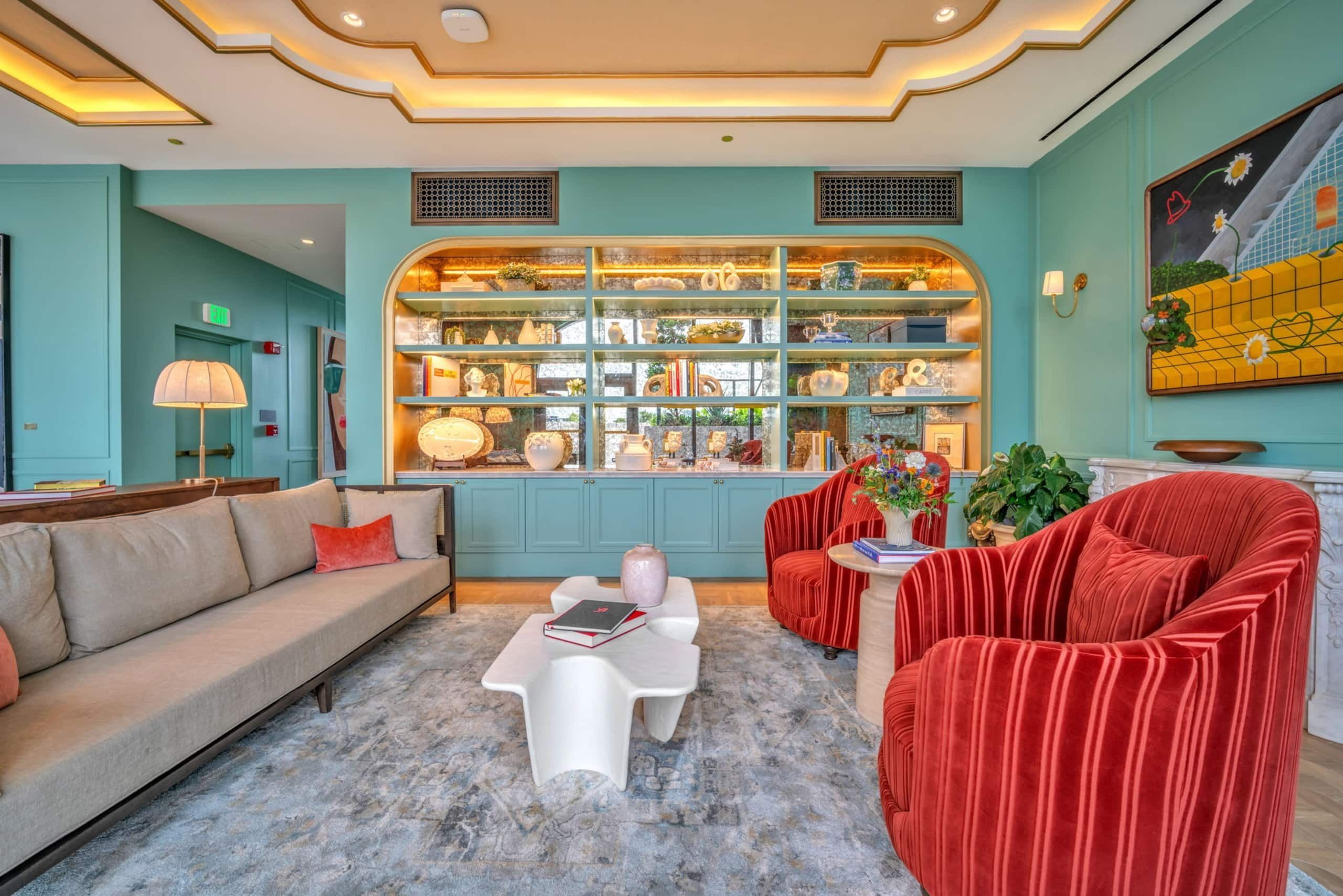 Un hall d’hôtel aux couleurs turquoise, avec un canapé, des fauteuils en velours rouge, une table basse et une étagère décorative où sont exposés divers objets.