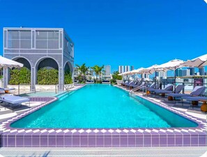 A pool area with lounge chairs and umbrellas, a clear blue sky, and a modern building with arched windows.