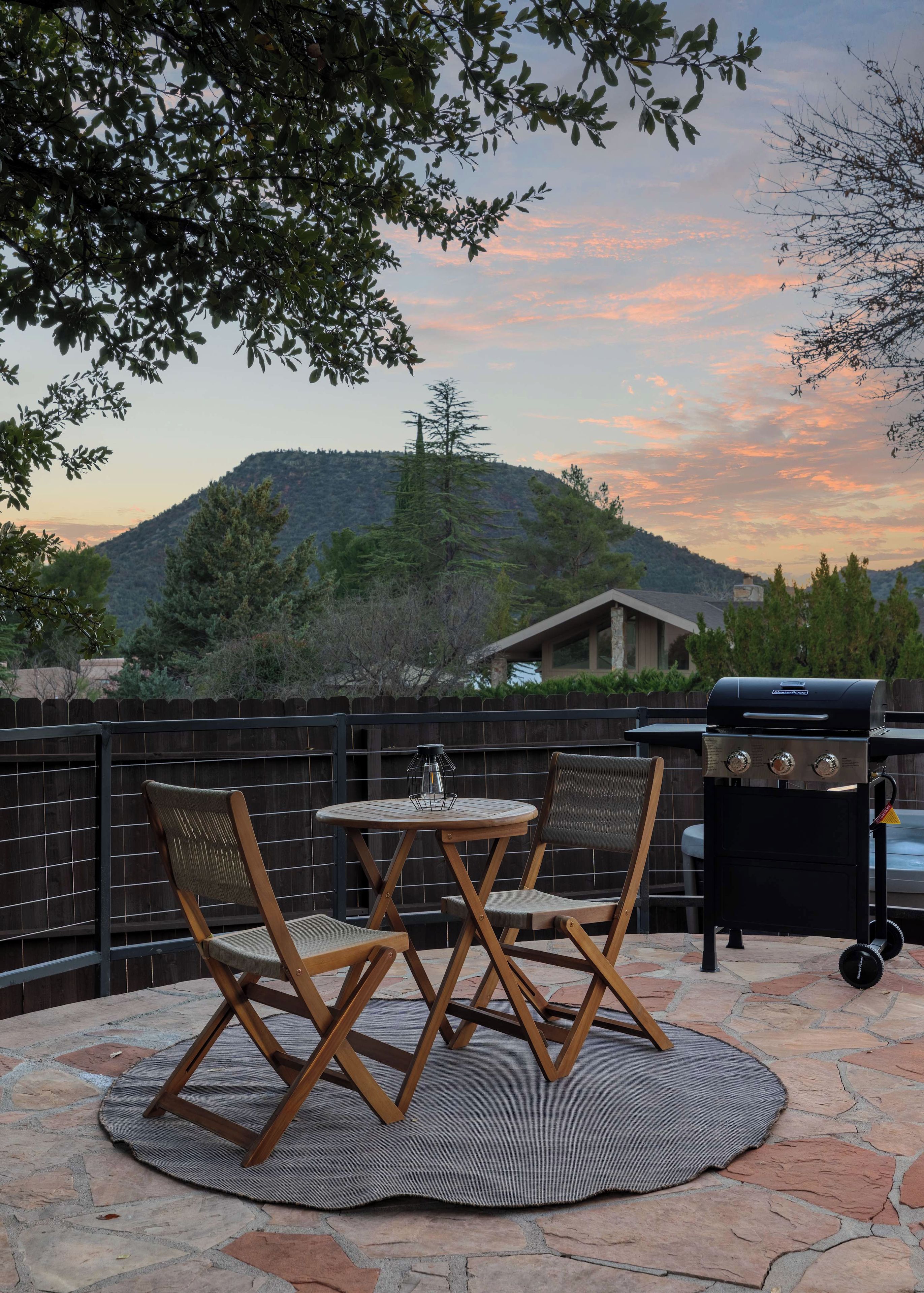 An outdoor patio with a round table and chairs, a grill, and a view of a hilly landscape.