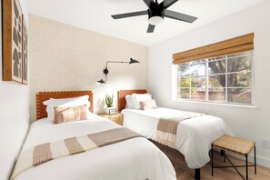 Ein Schlafzimmer mit zwei Betten, einem Deckenventilator, einem Nachttisch mit Lampe, einem Fenster mit Jalousien und Blick ins Freie.