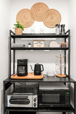 Eine Küchenzeile mit Kaffeemaschine, Mikrowelle und einem Regal, das gewebte Untersetzer, ein gerahmtes Bild und einen Topf mit Pflanze beherbergt.