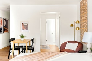 Ein Hotelzimmer mit einem Bett, einem Schreibtisch mit Stühlen, einem Esstisch, einem roten Sessel, einer Lampe und einer Wand mit geometrischem Muster.