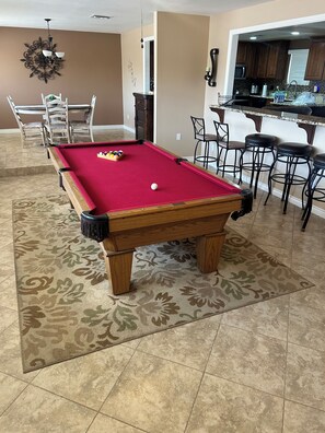 A pool table with a red felt surface, surrounded by a patterned rug, in a room with a kitchen area visible in the background. -  Snowbird Friendly. Monthly Discount
Delightful 4-bedroom home in Bullhead City. (Bullhead City)