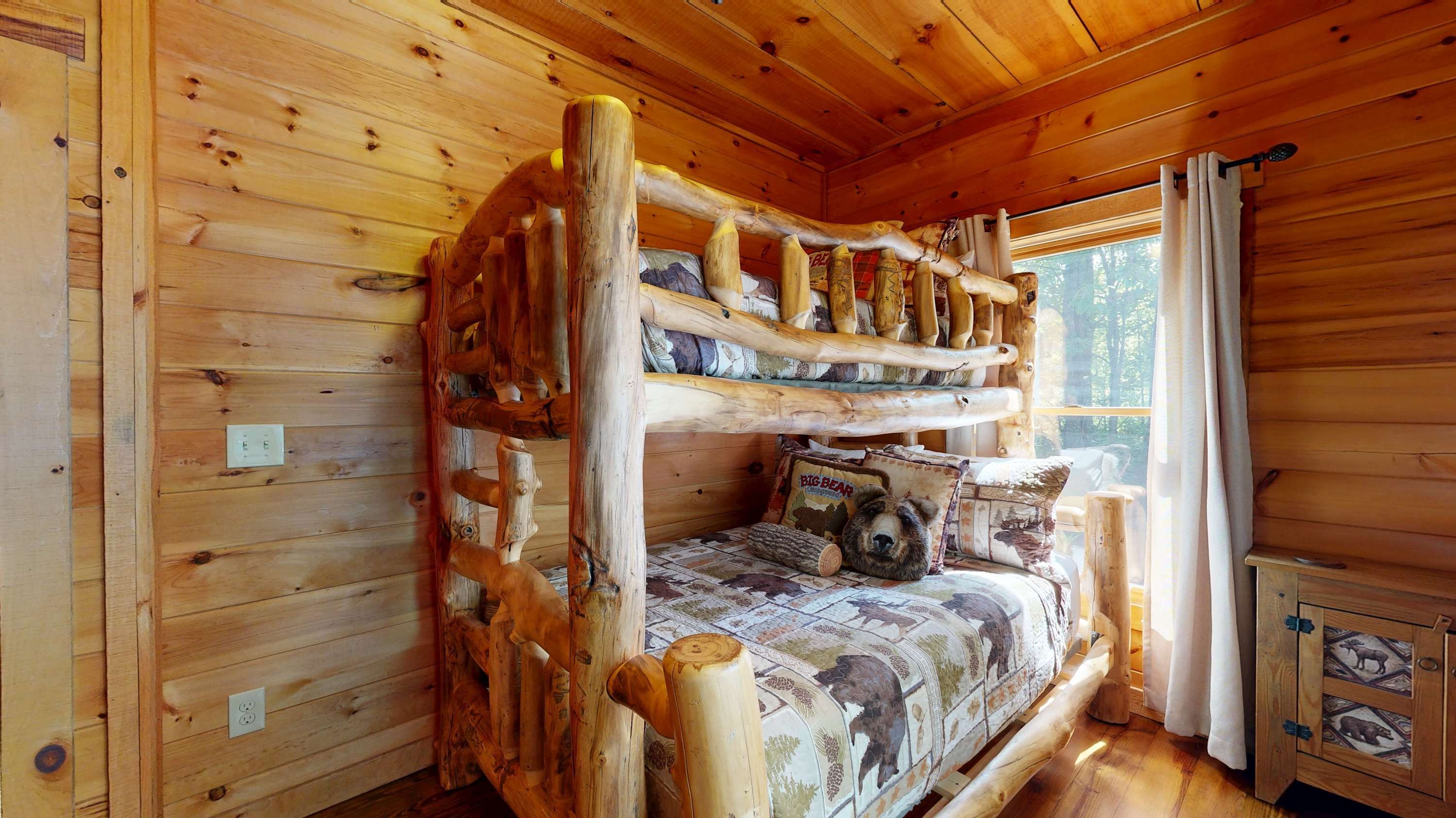 A bunk bed with a wooden frame and a patterned bedspread, a window with curtains, and a small wooden cabinet with animal designs.