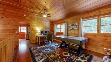 A wooden cabin interior with a pool table, a desk with a computer, and a ceiling fan.