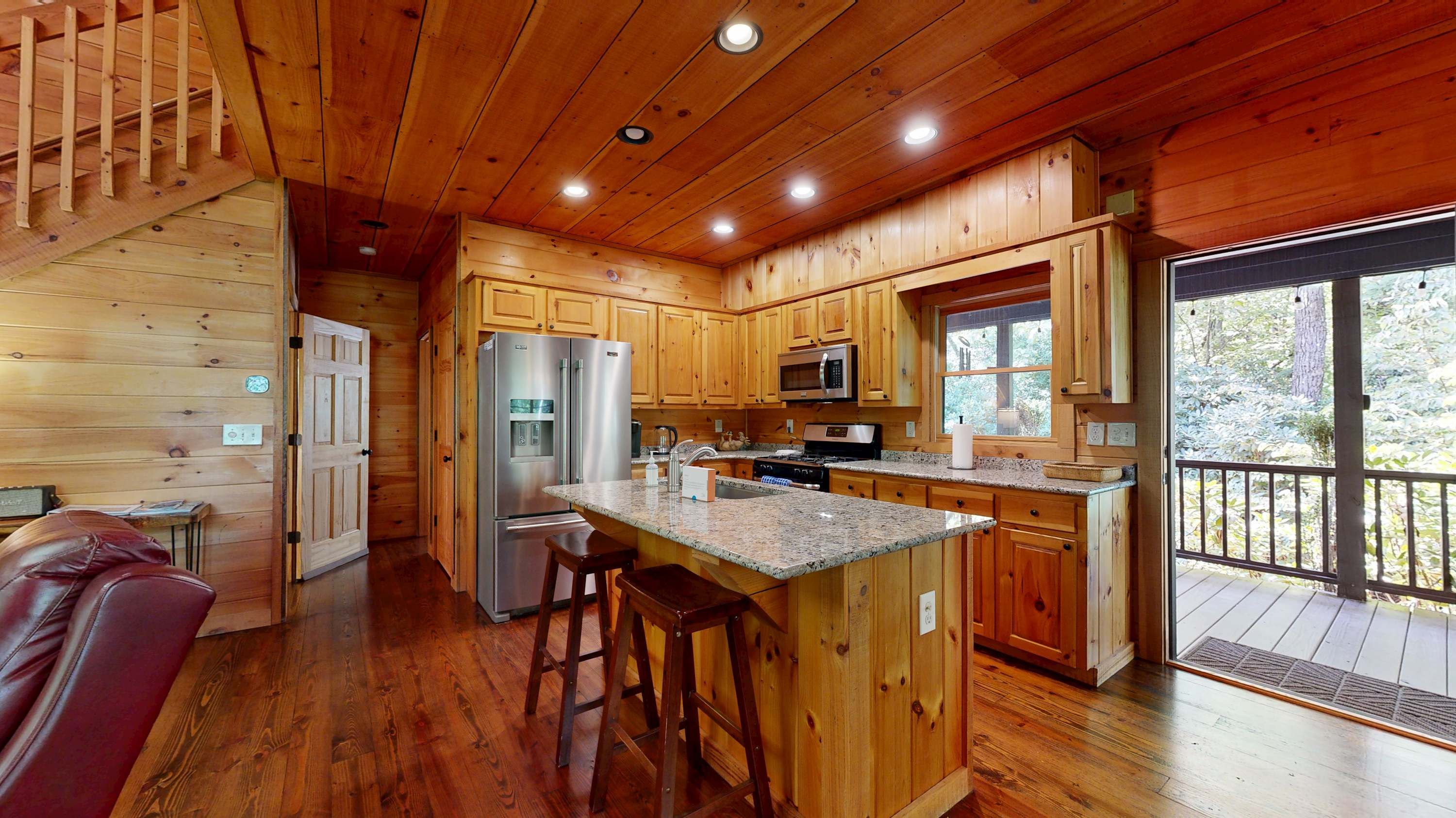 A cabin interior with wooden walls, a kitchen with granite countertops, a refrigerator, and a large window.
