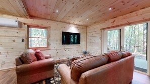 A log cabin interior with a television mounted on the wall, leather sofas, and large windows offering a view of the forest. - Thunder Mountain Lodge (Blue Ridge)