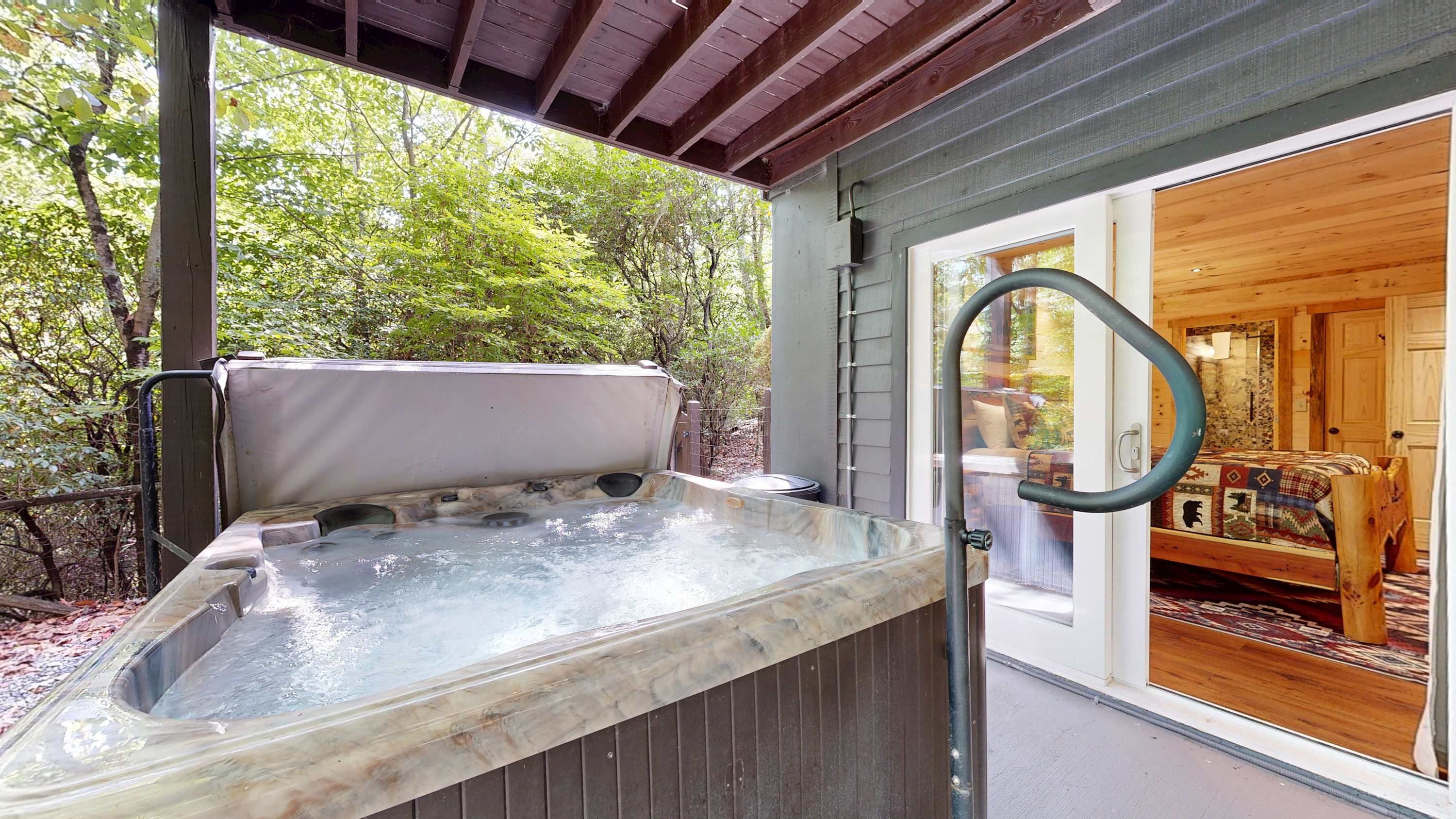 A hot tub with a wooden bench, surrounded by a wooden deck and a glass door leading to a room with a bed and a patterned rug.