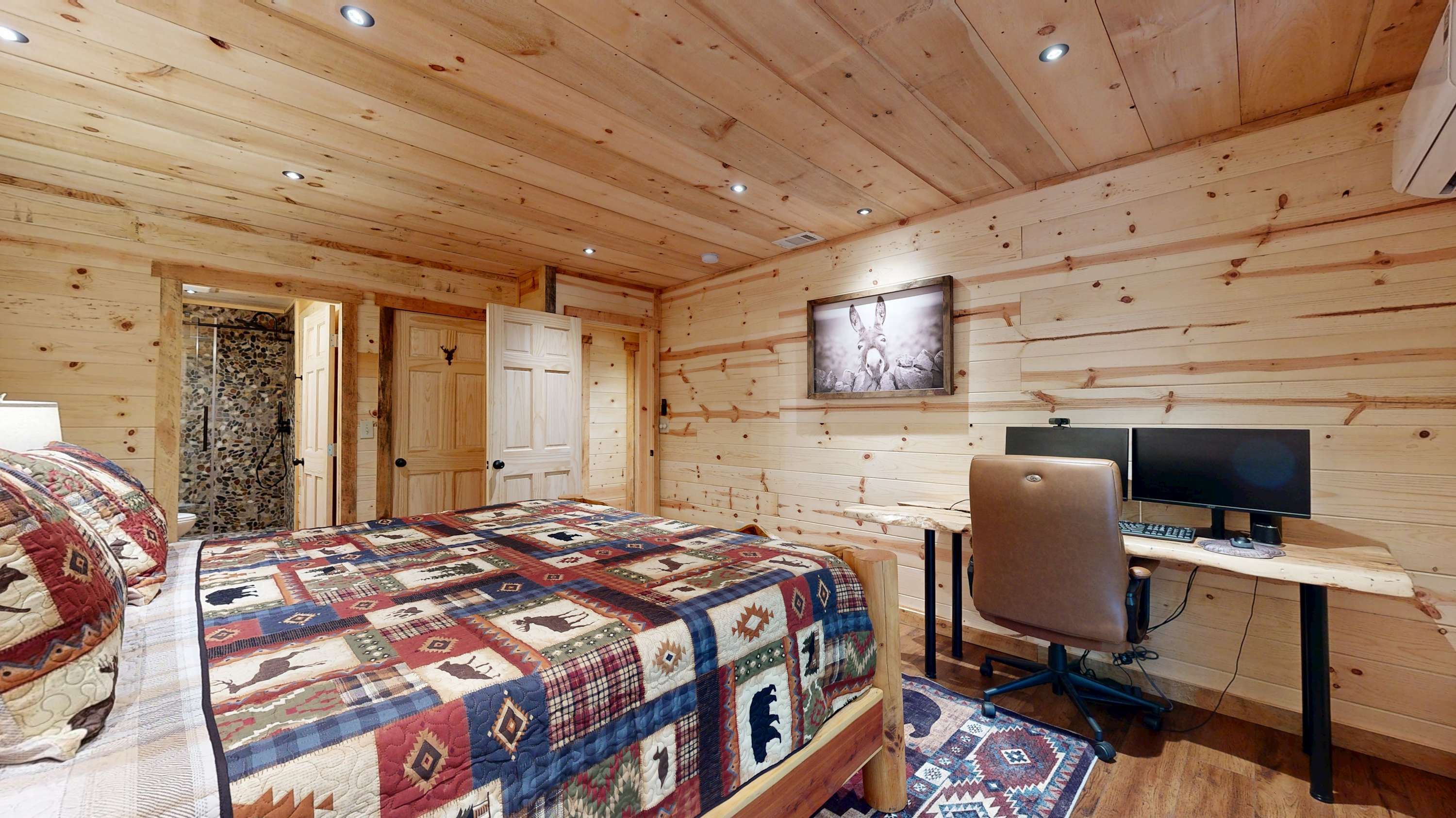 A wooden cabin bedroom with a bed, a desk with a computer, and a shower area.