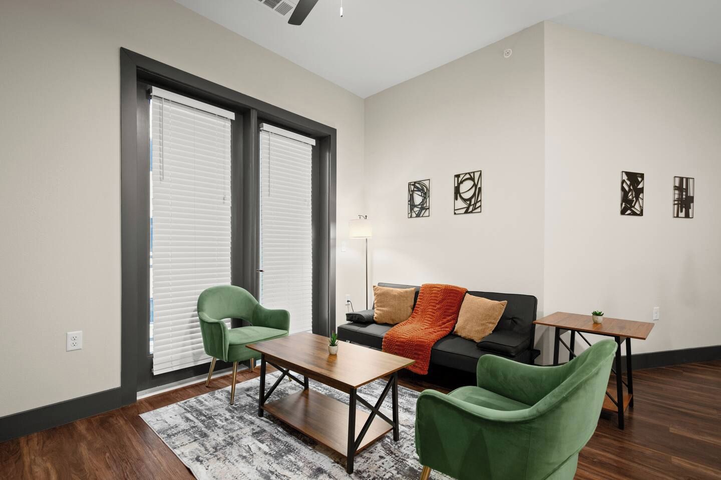 Een moderne woonkamer met een bank, twee fauteuils, een salontafel en een bijzettafeltje. Twee grote ramen met zonwering, een vloerlamp en abstracte wanddecoratie.