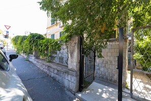 A gated residential area with a paved road, a car parked on the side, and a building with a stone wall and a metal gate. - Bartulin residence (Split)