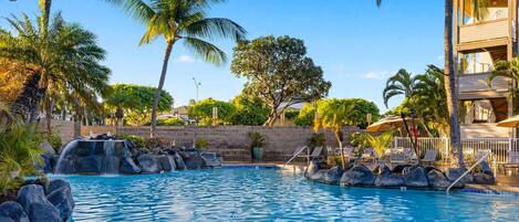 A swimming pool with a waterfall feature, surrounded by palm trees and lounge chairs.