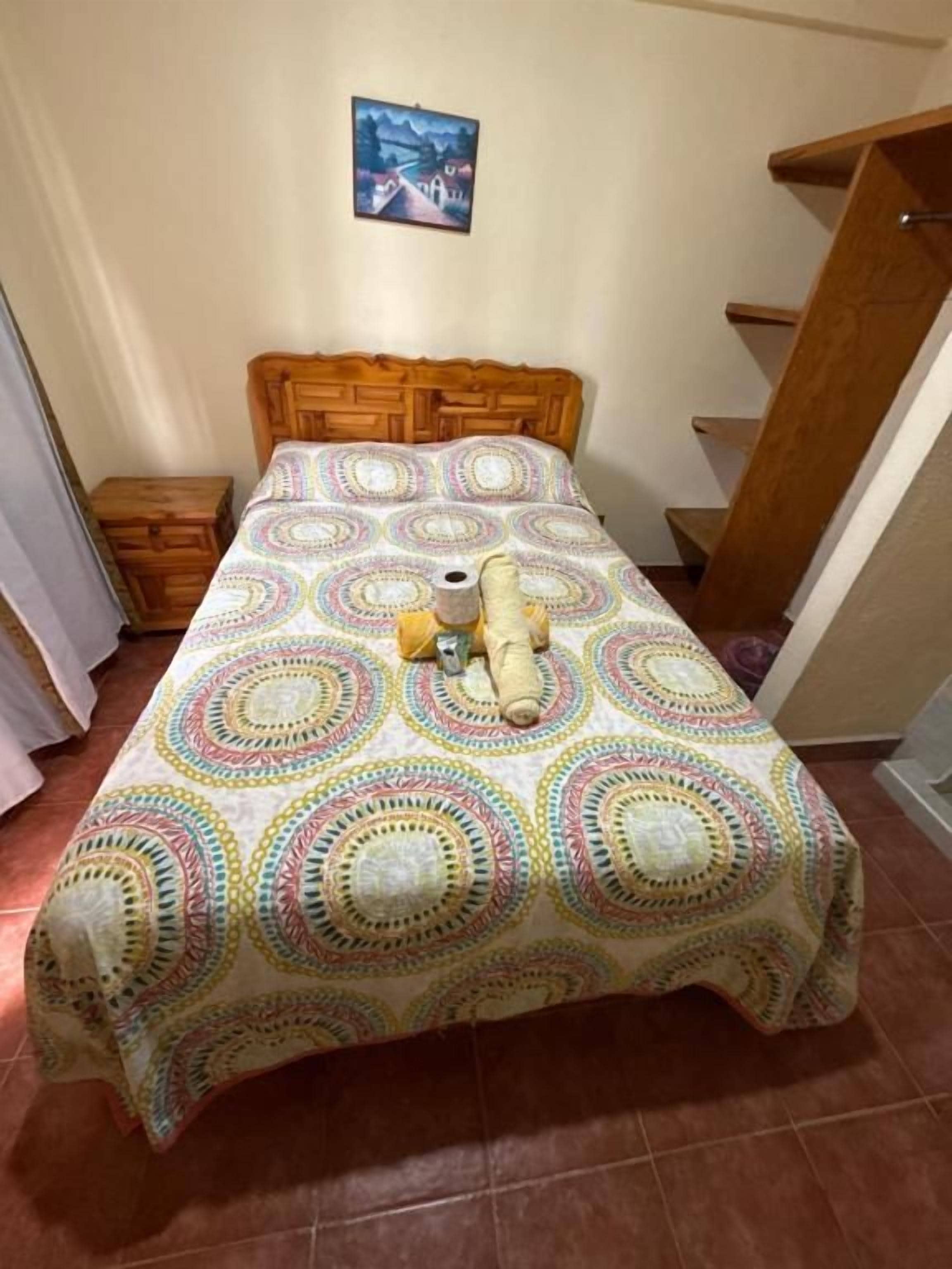 Una cama con colcha estampada, cabecero de madera, mesita de noche y un cuadro colgado en la pared.