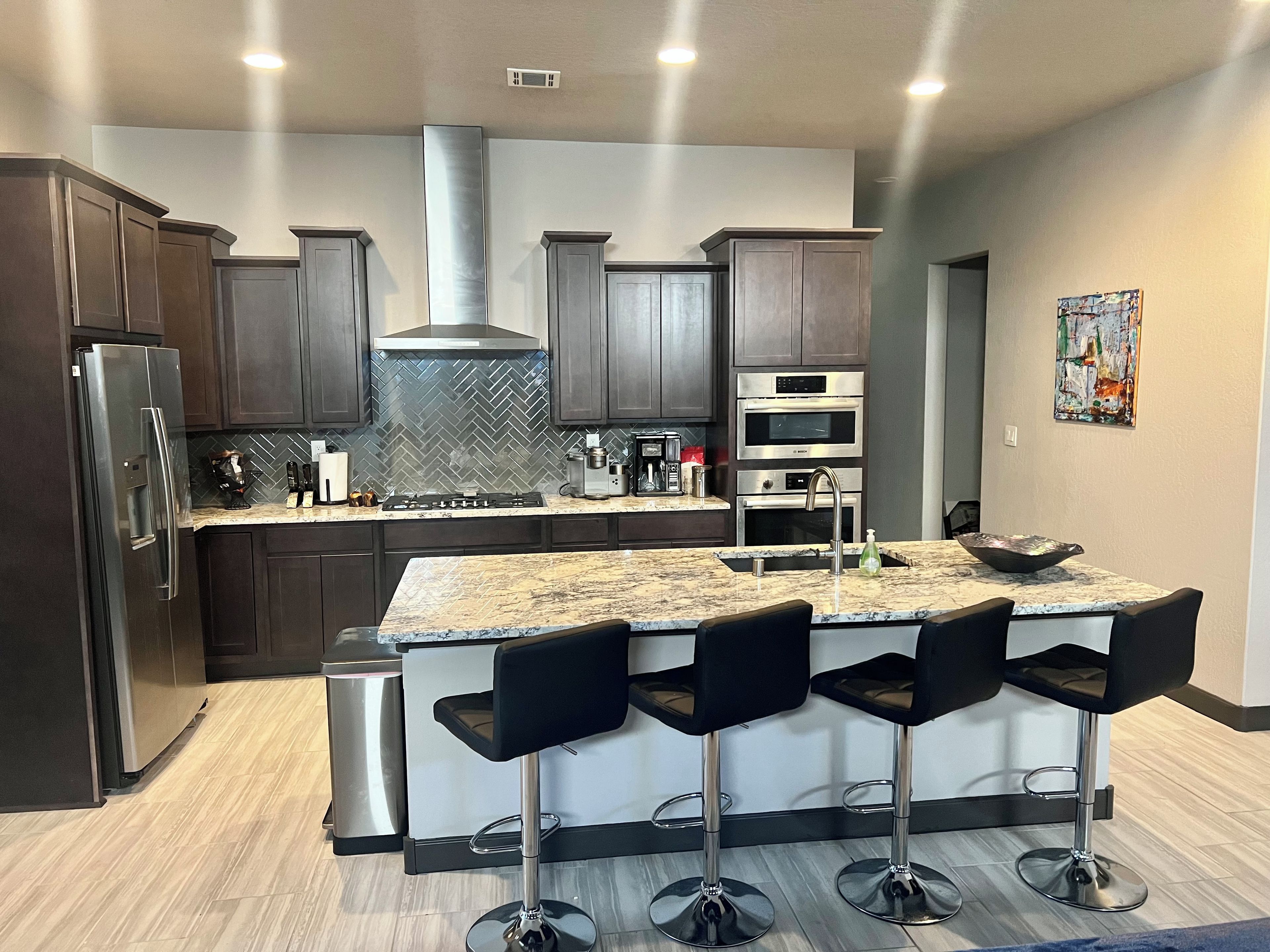 A modern kitchen with dark wood cabinets, stainless steel appliances, a herringbone backsplash, and a central island with bar stools.