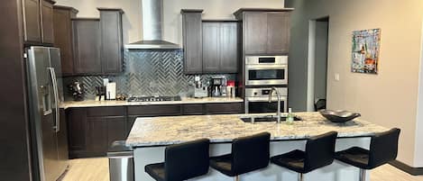 A modern kitchen with dark wood cabinets, stainless steel appliances, a herringbone backsplash, and a central island with bar stools.