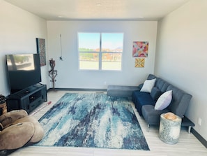 A living room with a flat-screen TV, a grey sofa, a patterned rug, and a window with a view of buildings. - Brilliant, Modern 3-bedroom house with AC in vibrant Albuquerque (Albuquerque)