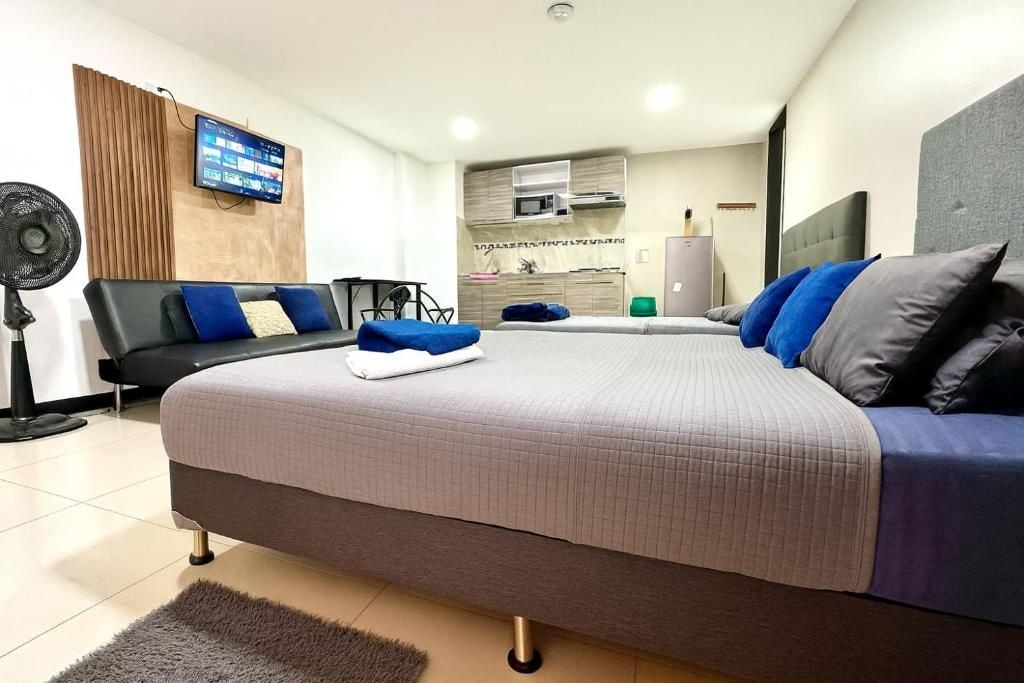 A modern bedroom with a large bed, a flat-screen TV, a desk, and a kitchenette in the background.