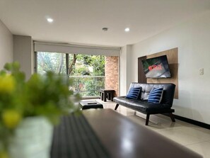 A modern living room with a leather sofa, a flat-screen TV mounted on the wall, and a large window providing a view of greenery outside. - Tolupa Apartamentos (Medellín)